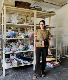 Artist I. Ronni Kappos stands smiling in front of some shelves that contain bags of colorful beads.